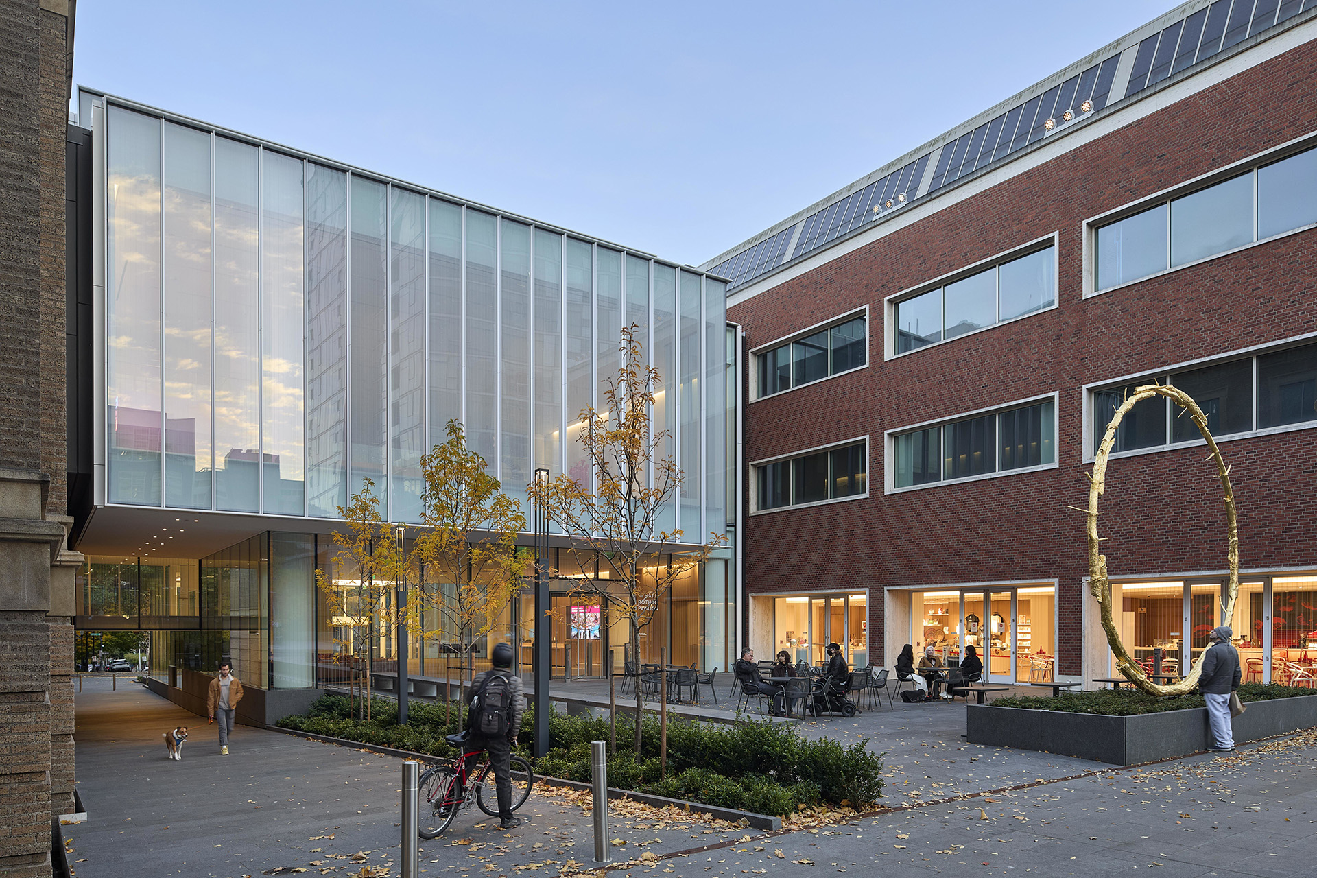 Modern glass structure blends with red brick buildings, featuring trees and a circular sculpture, with people and a dog in the plaza.