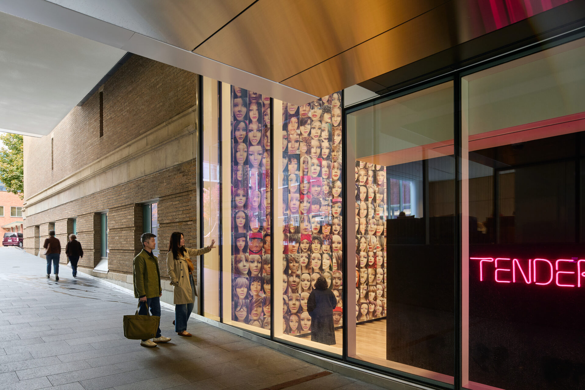 Two visitors interact with a colorful mural of diverse faces displayed in a large glass window, alongside glowing neon text reading "TENDER."