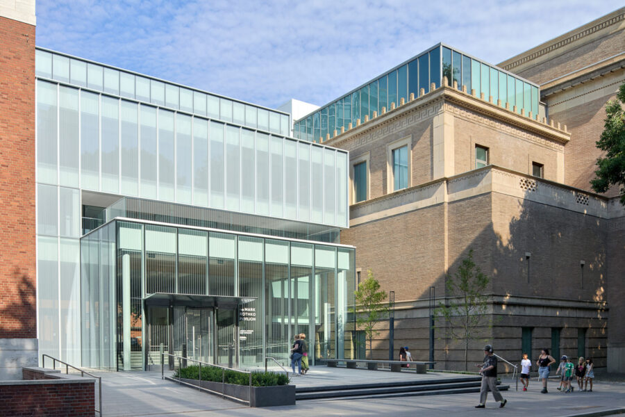 Modern glass entryway leading to a museum, flanked by brick and stone buildings, with trees and people in a well-lit outdoor space.