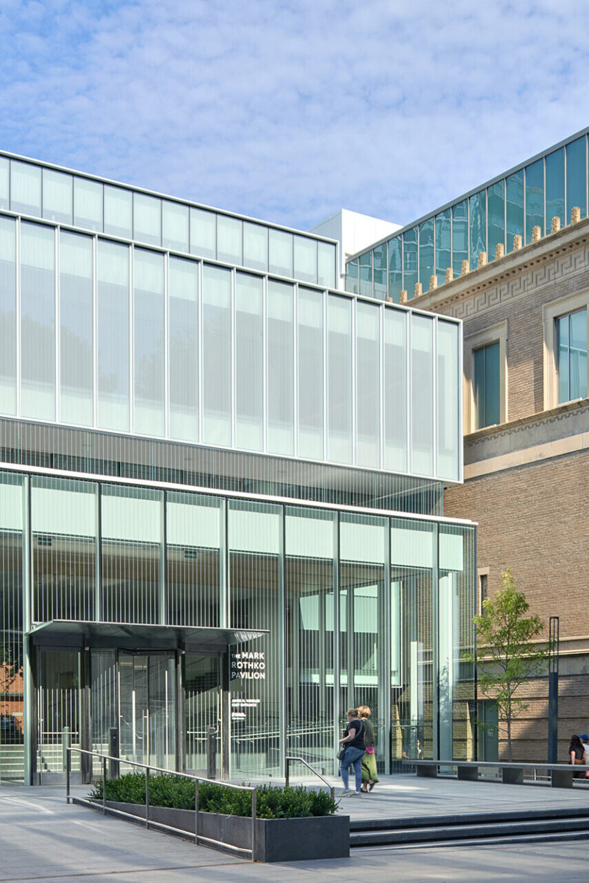 Modern glass and metal building with a green accent, featuring wide steps, a plaza, and visitors walking underneath a cloudy sky.
