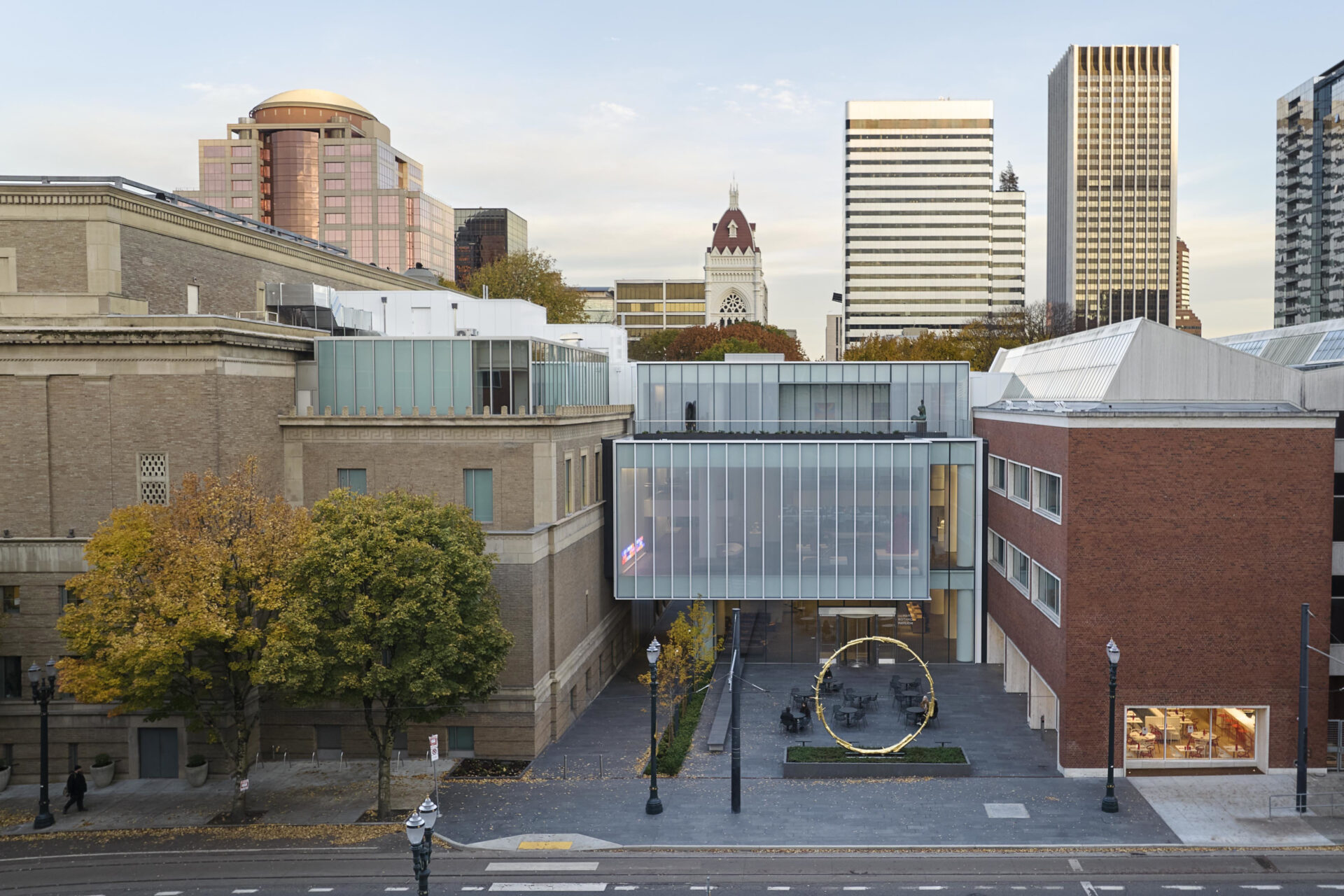 Modern glass pavilion of the Portland Art Museum integrates with urban landscape, surrounded by trees and tall city buildings under a soft sky.