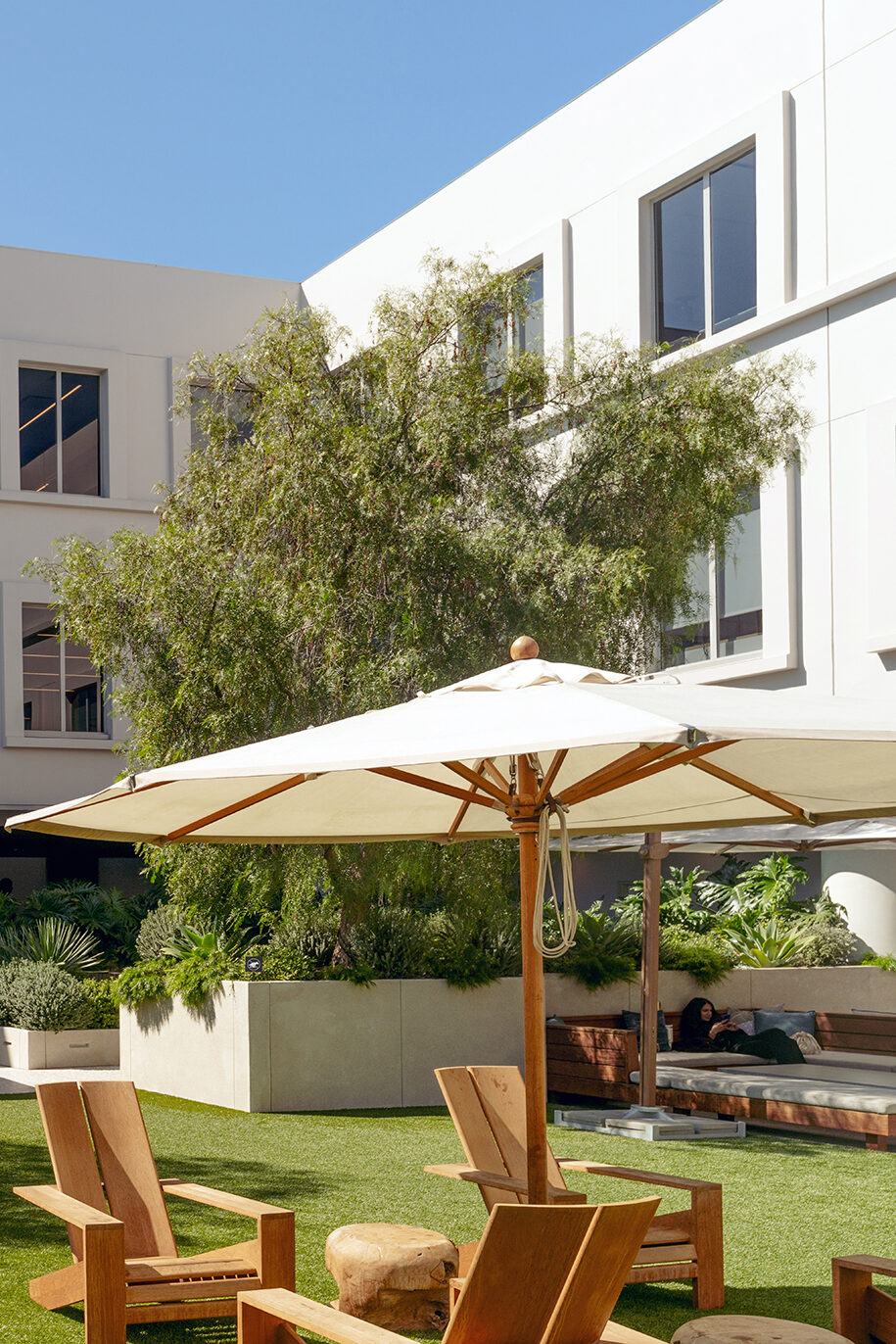 A serene outdoor space featuring wooden lounge chairs, a large white umbrella, lush greenery, and a modern building backdrop.