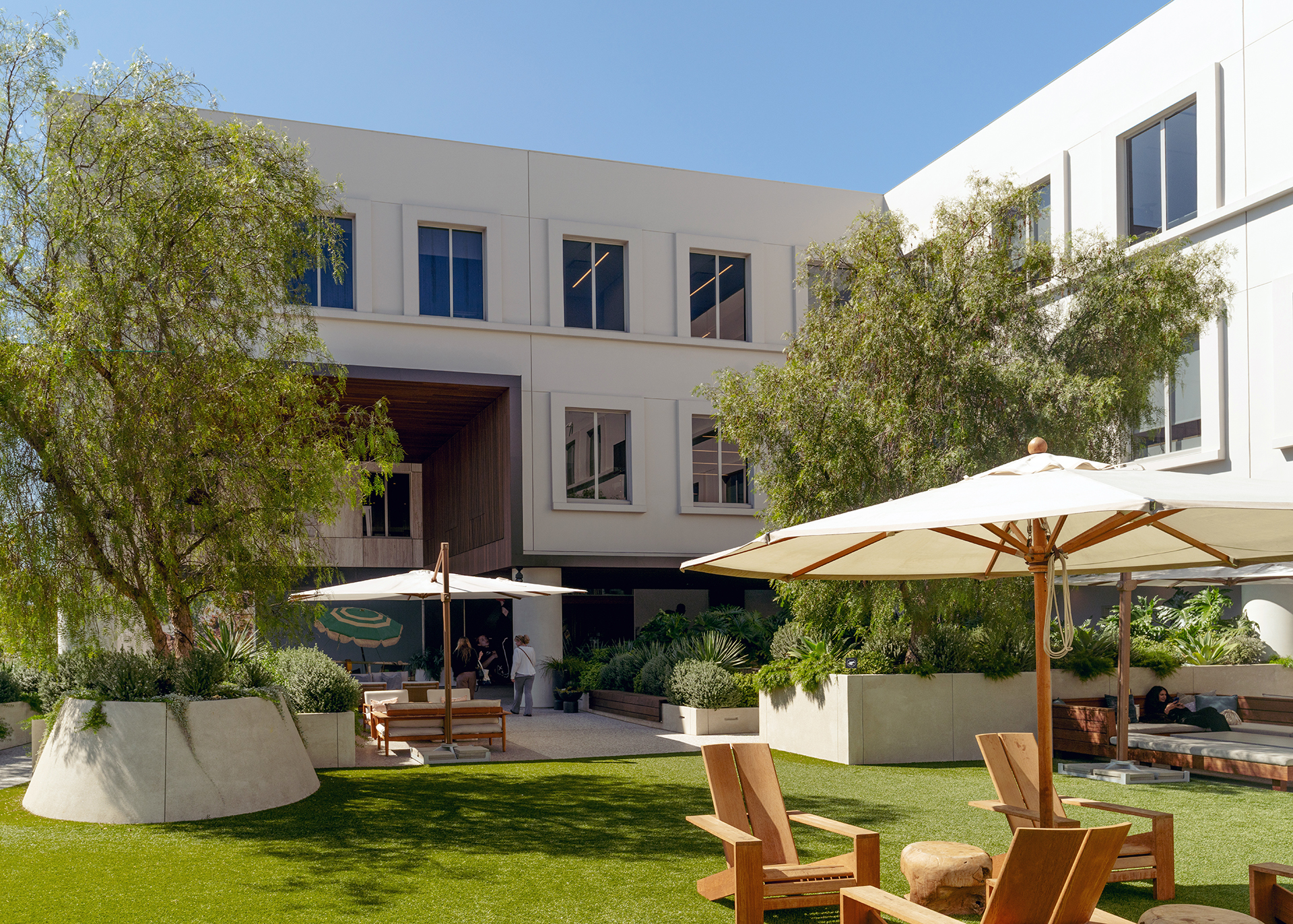 A modern courtyard with greenery, wooden loungers, and umbrellas under a clear blue sky, flanked by a contemporary building.