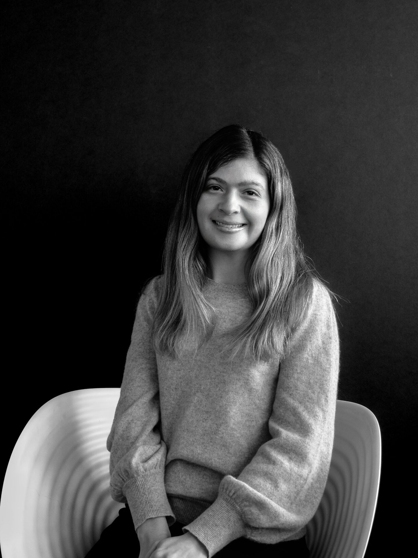 A woman with long hair, wearing a light gray sweater, sits on a white chair against a dark background, smiling confidently.