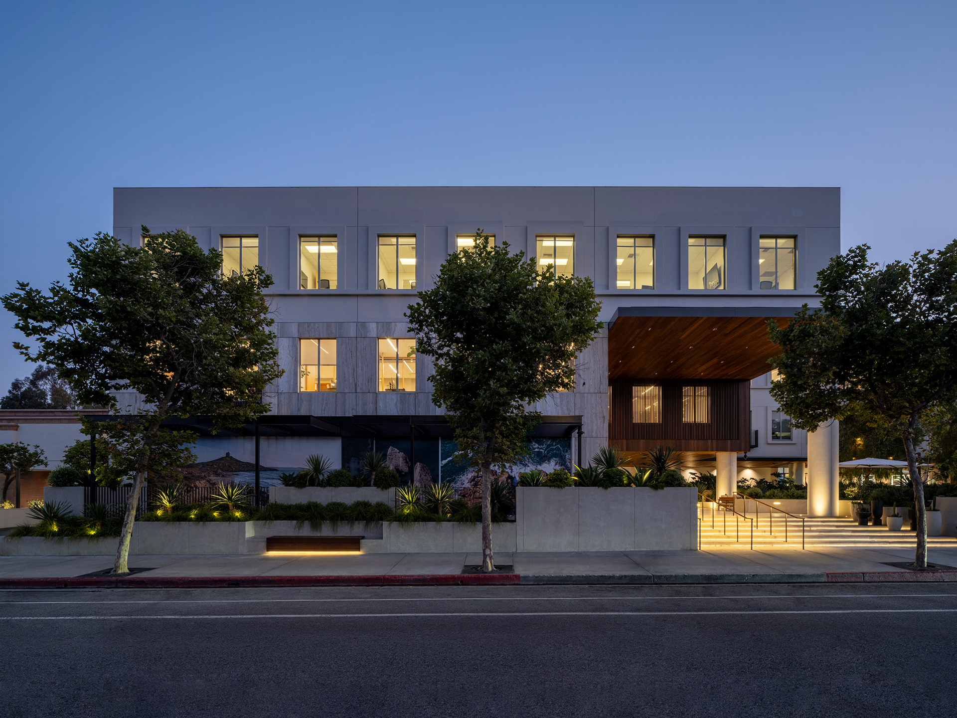 Modern building facade at dusk, featuring large windows, wooden accents, and landscaped greenery, creating an inviting atmosphere.