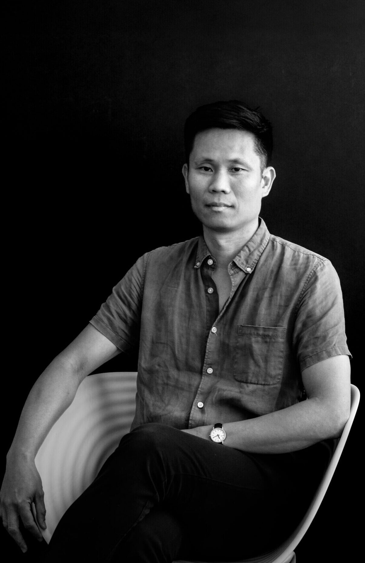 Ariel Dungca, dressed in a casual button-up shirt, sits confidently in a modern chair against a black background.