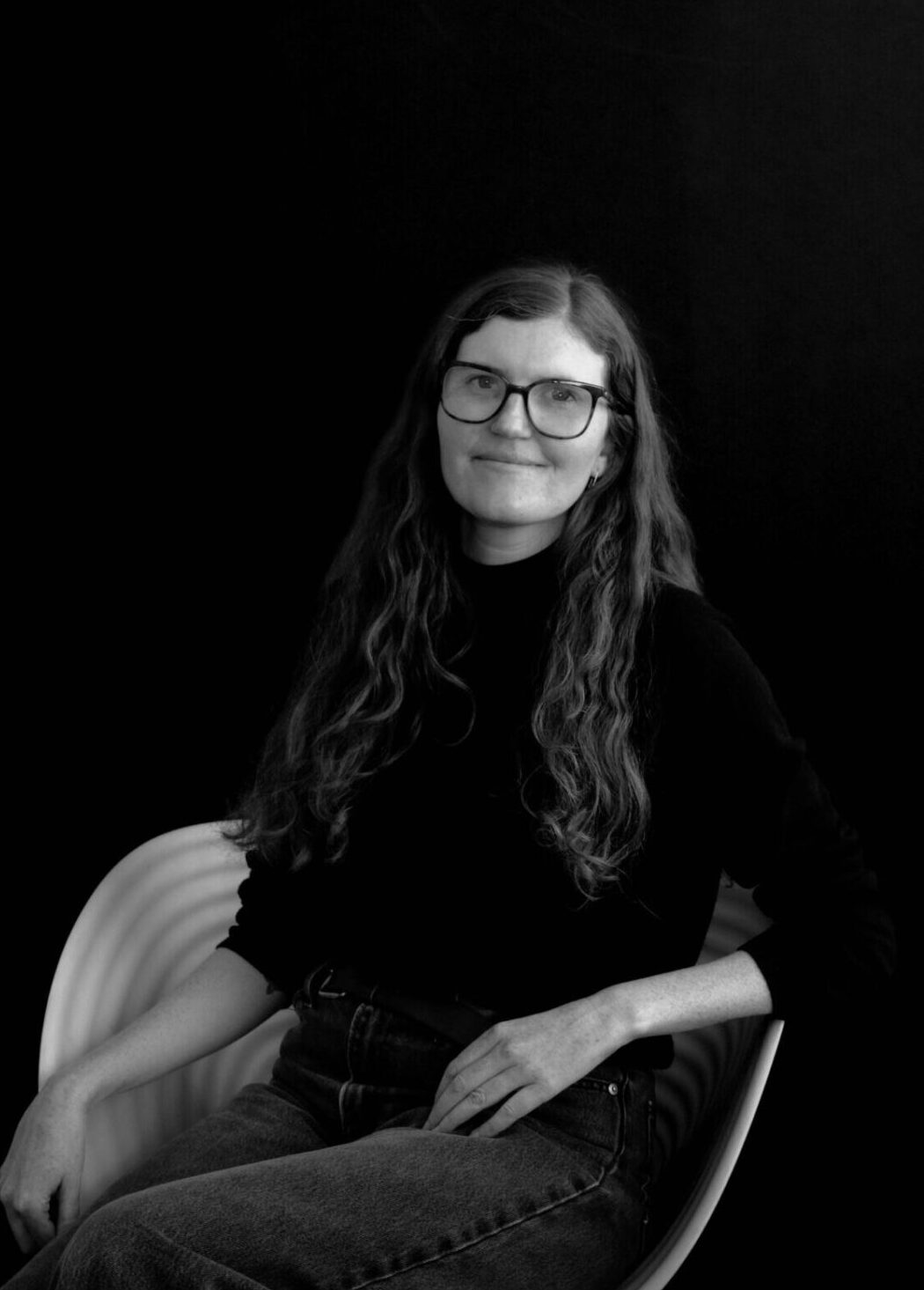 A smiling woman with long hair and glasses sits on a white chair against a black background, wearing a black turtleneck and jeans.